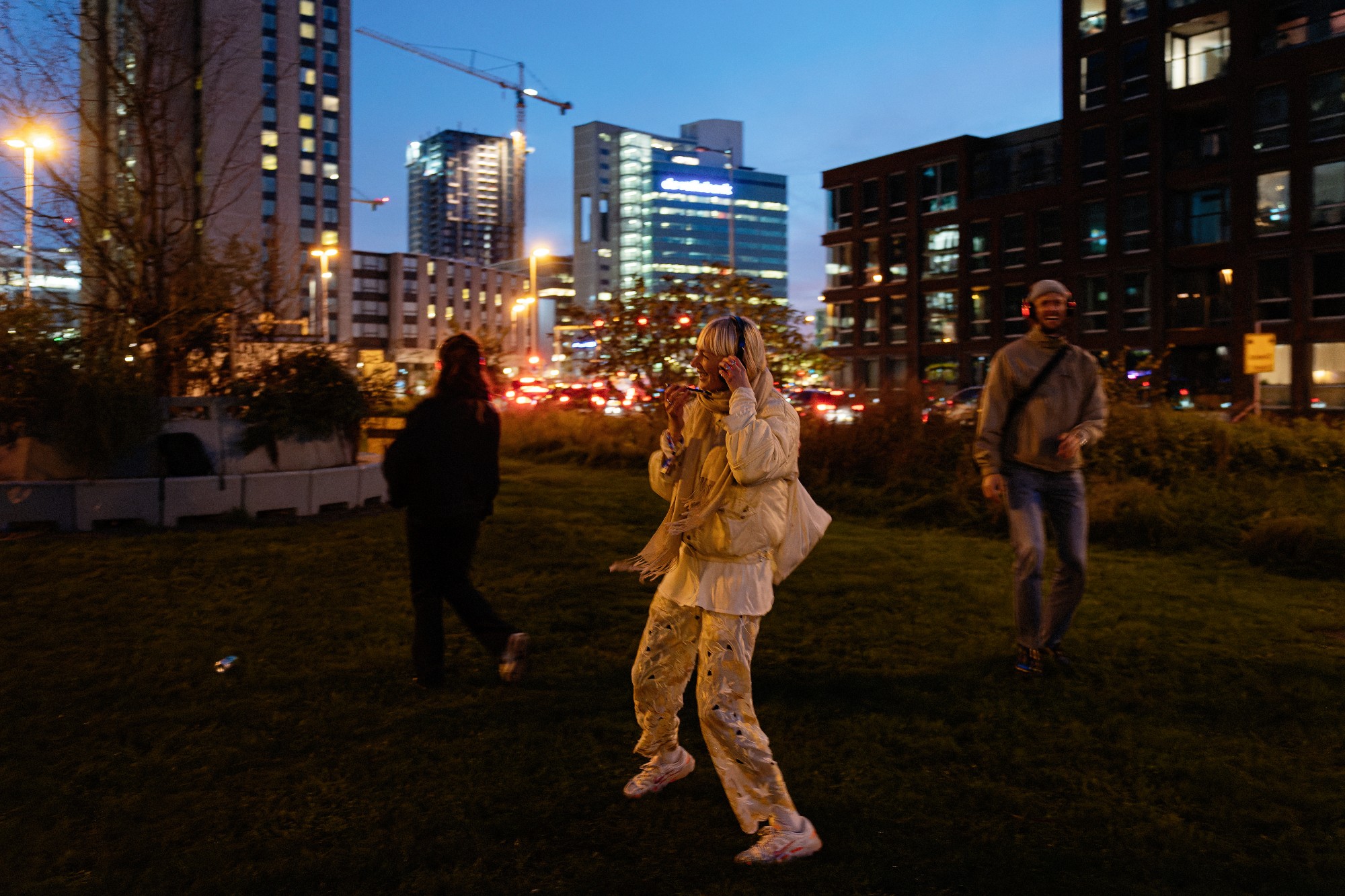 A person fully dressed in beige is dancing, wearing headphones, in the front of the photo. Two other people are behind, with some modern illuminated building in the background